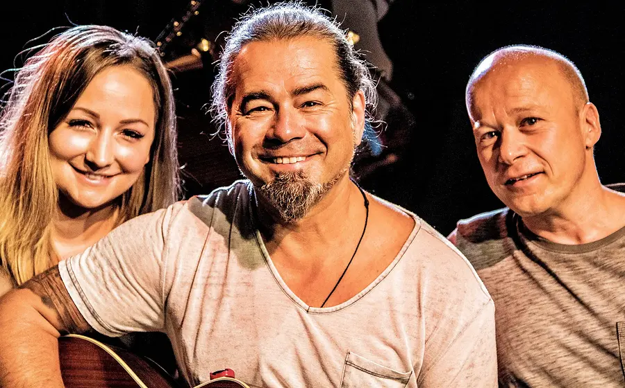 Eine Dame und zwei Herren in Freizeitkleidung, einer davon mit einer Gitarre in der Hand, vor dunklem Hintergrund