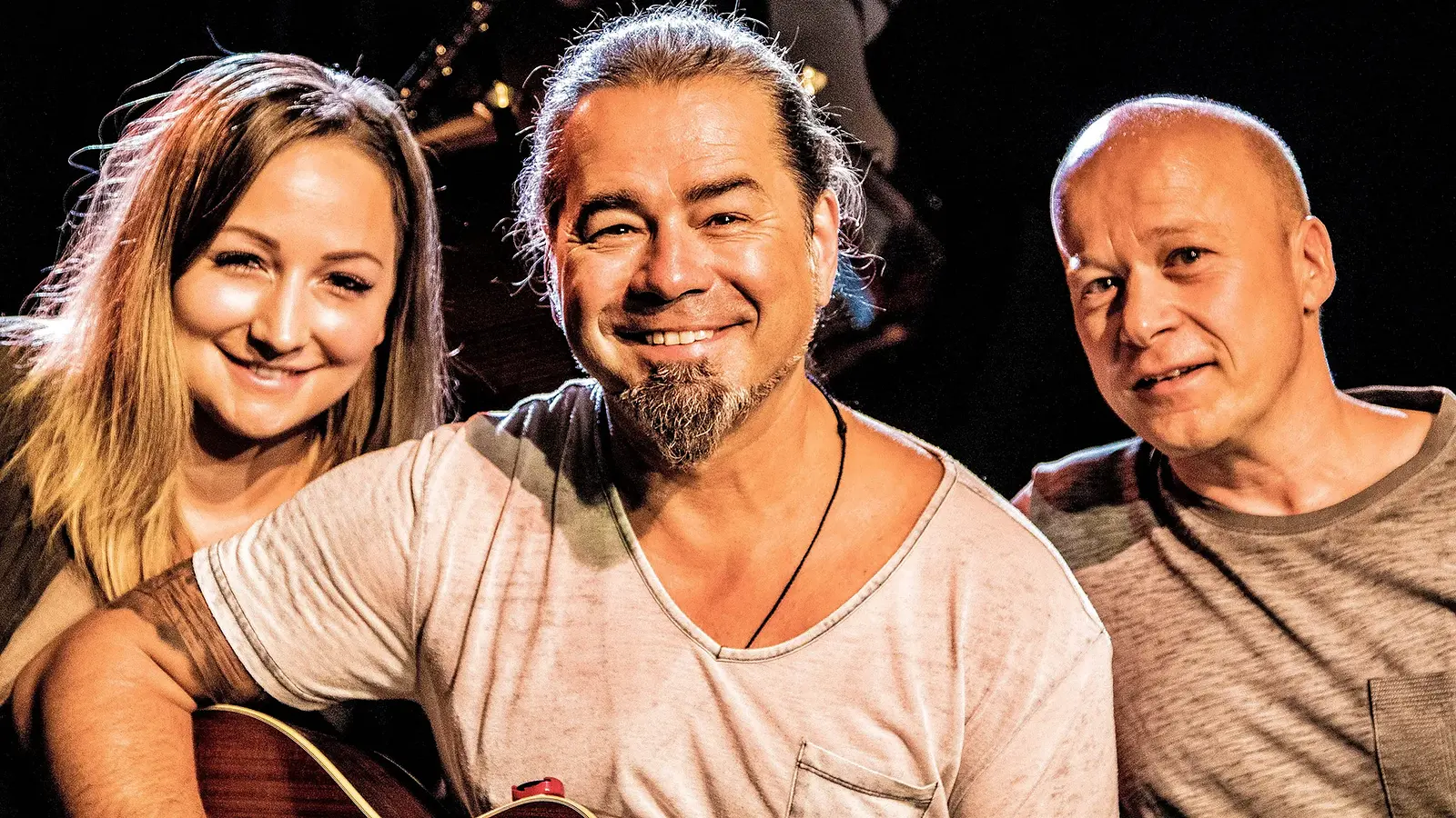 Eine Dame und zwei Herren in Freizeitkleidung, einer davon mit einer Gitarre in der Hand, vor dunklem Hintergrund