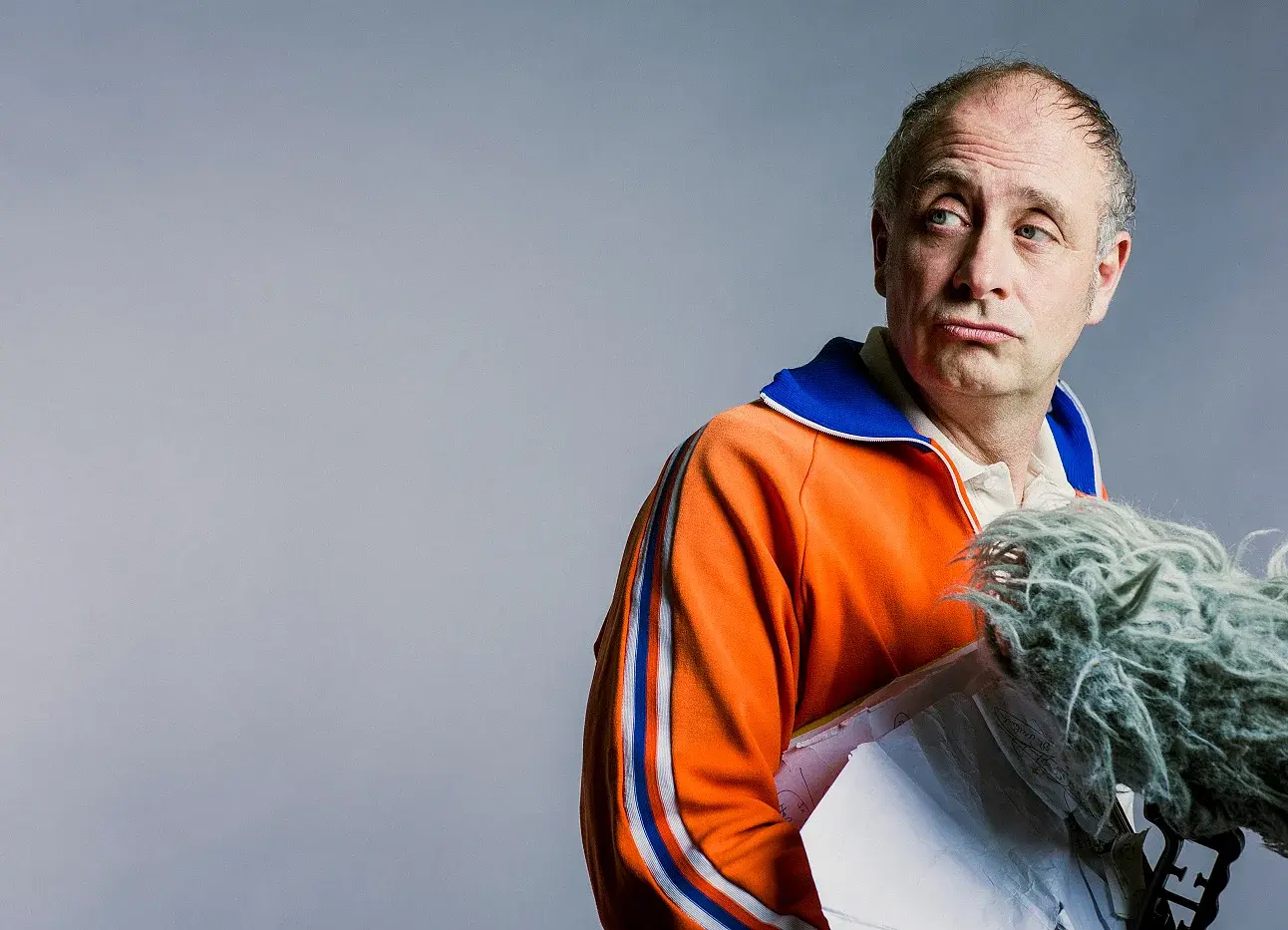 Foto von Alfons in orangefarbener Trainingsjacke mit blauem Kragen über hellem Poloshirt, vor grauem Hintergrund, mit seitlichem Blick und einem Mikrofon mit Fellwindschutz im Vordergrund