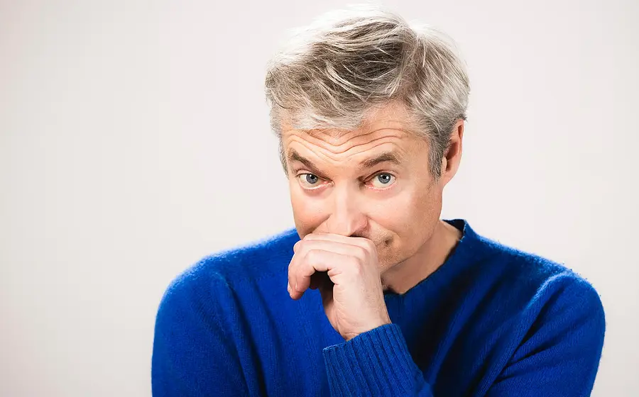 Portrait von Alfred Dorfer, der eine Hand vor den Mund hält, in blauem Pullover vor hellem Hintergrund