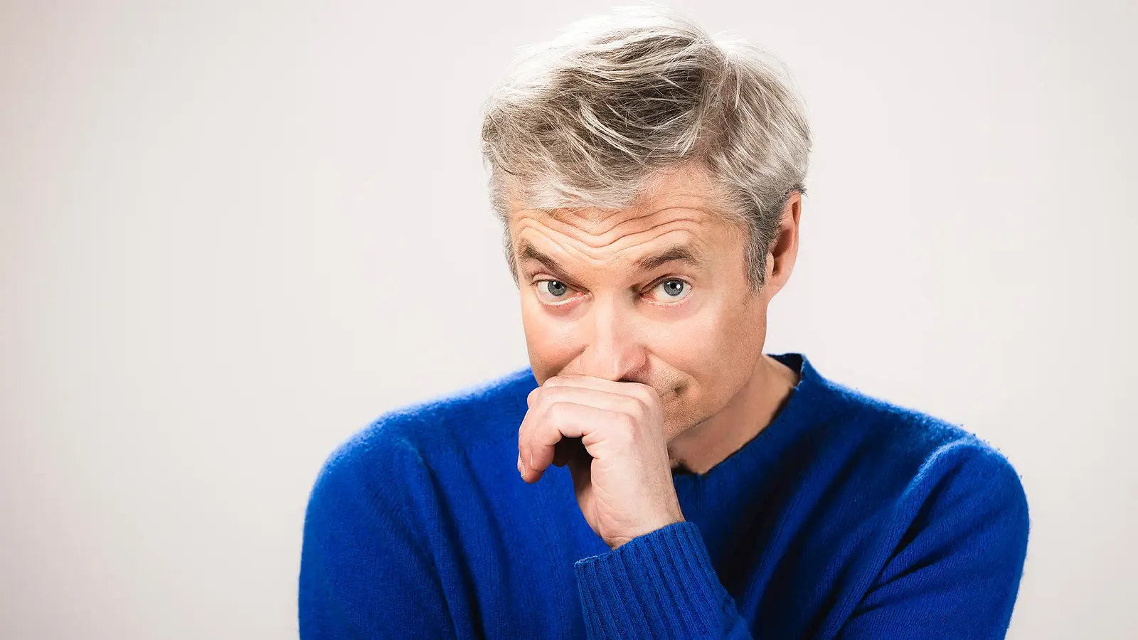 Portrait von Alfred Dorfer, der eine Hand vor den Mund hält, in blauem Pullover vor hellem Hintergrund