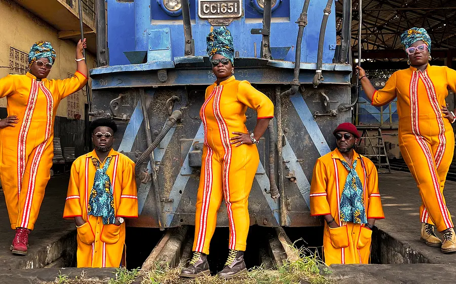 Gruppenfoto von fünf Personen in leuchtend orangefarbenen Kostümen vor einer blauen Lokomotive in einem Bahndepot. Die Personen tragen farbige Kopfbedeckungen und Sonnenbrillen und posieren symmetrisch vor der Lok auf den Gleisen.