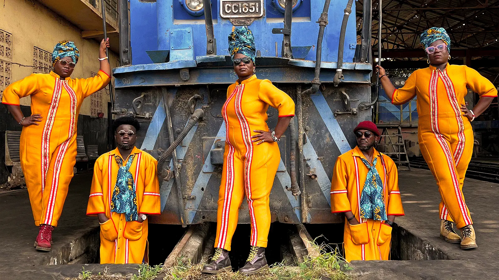 Gruppenfoto von fünf Personen in leuchtend orangefarbenen Kostümen vor einer blauen Lokomotive in einem Bahndepot. Die Personen tragen farbige Kopfbedeckungen und Sonnenbrillen und posieren symmetrisch vor der Lok auf den Gleisen.