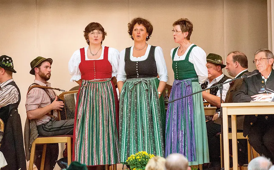 Drei Damen in Tracht, im Hintergrund weitere Musiker und Teilnehmer zur Veranstaltung Alpensingen