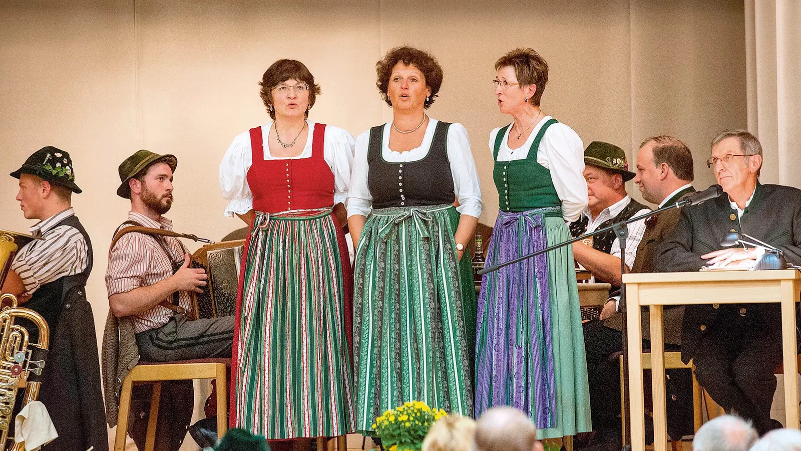Drei Damen in Tracht, im Hintergrund weitere Musiker und Teilnehmer zur Veranstaltung Alpensingen