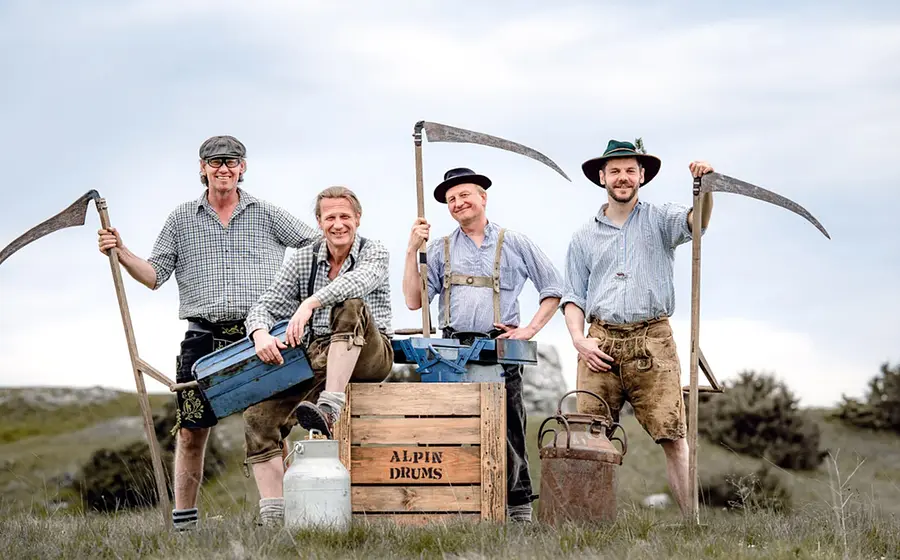 Vier Personen in traditioneller Arbeitskleidung stehen auf einer Wiese mit Sensen und einer Holzkiste mit der Aufschrift ‚Alpin Drums‘ vor ländlicher Kulisse