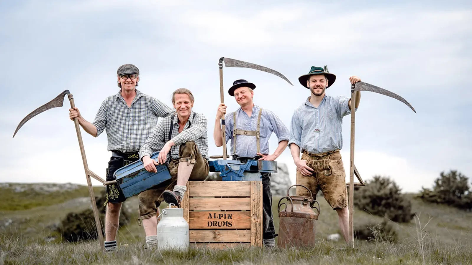 Vier Personen in traditioneller Arbeitskleidung stehen auf einer Wiese mit Sensen und einer Holzkiste mit der Aufschrift ‚Alpin Drums‘ vor ländlicher Kulisse