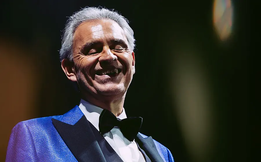 Andrea Bocelli in einem blauen Anzug mit weißem Hemd und schwarzer Fliege lachend vor dunklem Hintergrund mit Lichtreflexen