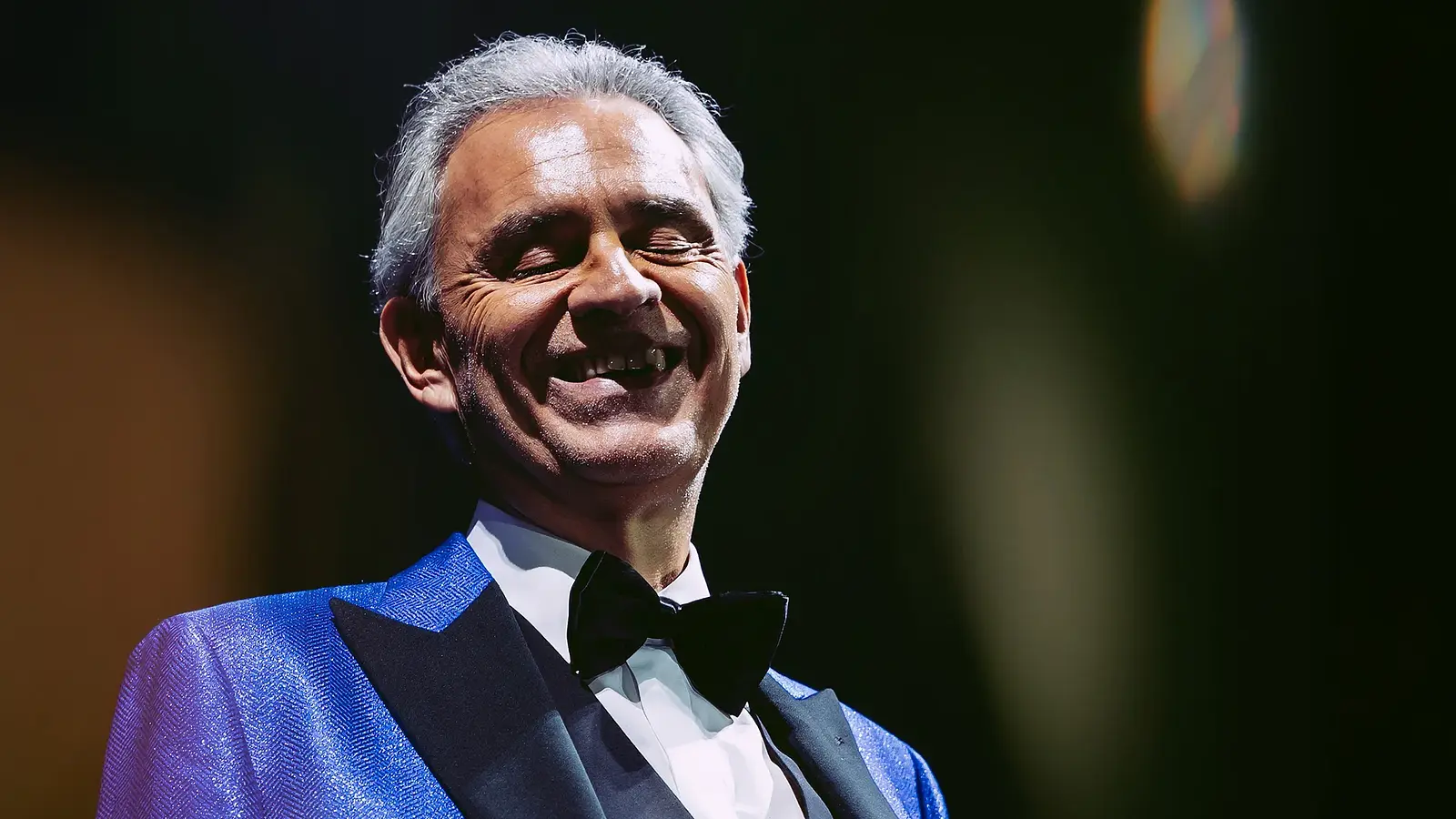 Andrea Bocelli in einem blauen Anzug mit weißem Hemd und schwarzer Fliege lachend vor dunklem Hintergrund mit Lichtreflexen