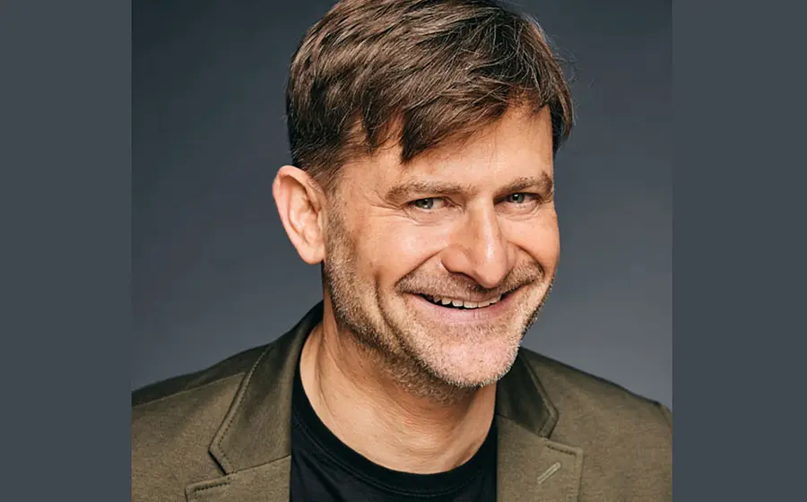 Michael Altinger lachend in olivgrünem Sakko und schwarzem T Shirt vor dunkelgrauem Hintergrund