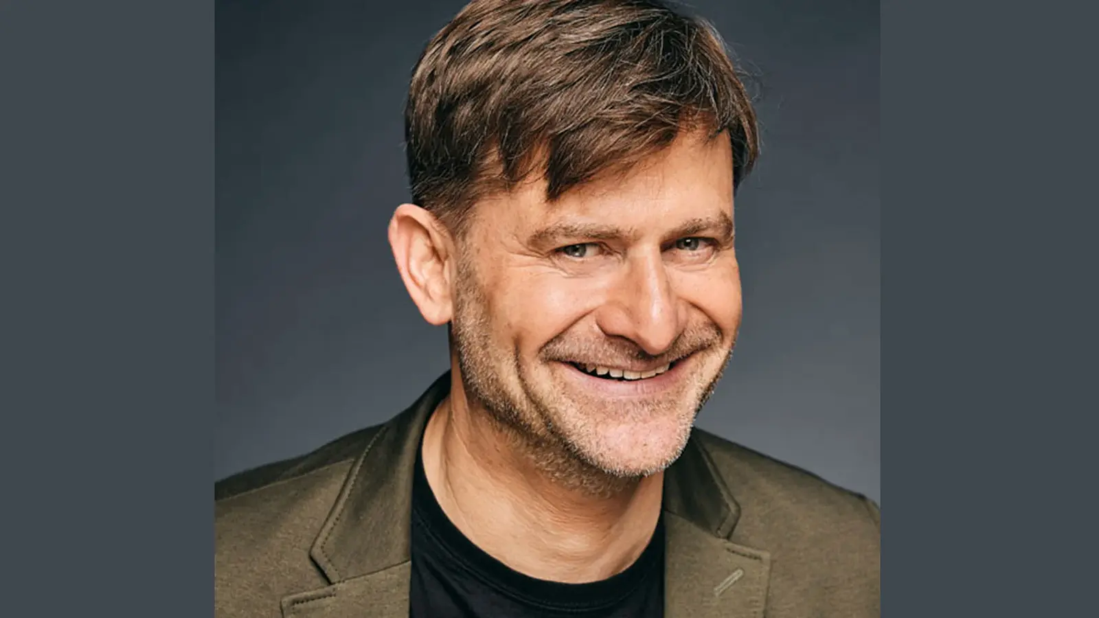 Michael Altinger lachend in olivgrünem Sakko und schwarzem T Shirt vor dunkelgrauem Hintergrund
