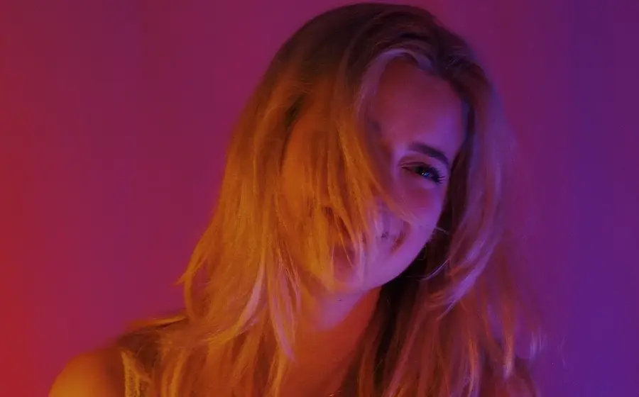 Porträtfoto von Amelie Scheffels mit langen blonden Haaren vor pink violett beleuchtetem Hintergrund, in Nahaufnahme mit seitlich ins Gesicht fallenden Haarsträhnen.