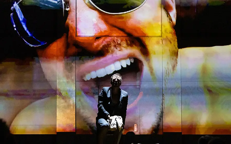 Bühnenszene mit einem sitzenden Mann im Vordergrund, hinter ihm eine große farbige Projektion eines Gesichts mit Sonnenbrille – Theatermotiv.