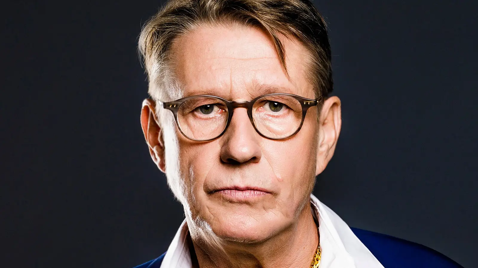 Ein Portrait von Andreas Rebers in weißem Hemd, goldener Kette und blauem Sakko vor dunklem Hintergrund