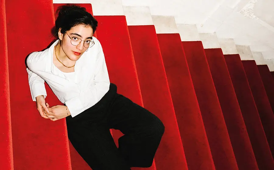 Eine Person sitzt auf einer roten, mit Teppich belegten Treppe. Sie trägt eine weiße Bluse, eine schwarze Hose und eine Brille. Die Treppe verläuft diagonal durch das Bild, der obere Bereich ist in hellen Weiß- und Cremetönen gestaltet.