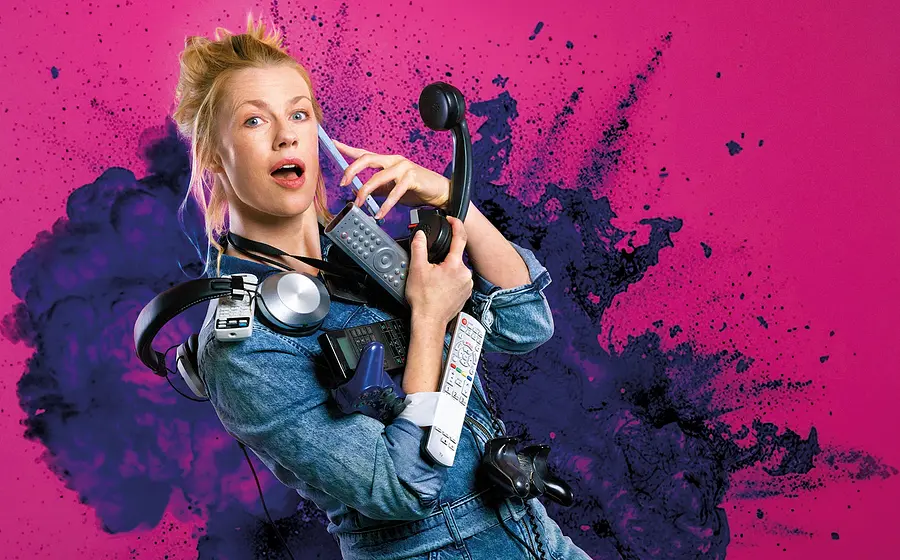 Christine Eixenberger im Jeansoutfit, die zahlreiche Alltagsgeräte wie Telefon Kopfhörer und Fernbedienungen hält, vor pink violettem Hintergrund mit Farbspritzern