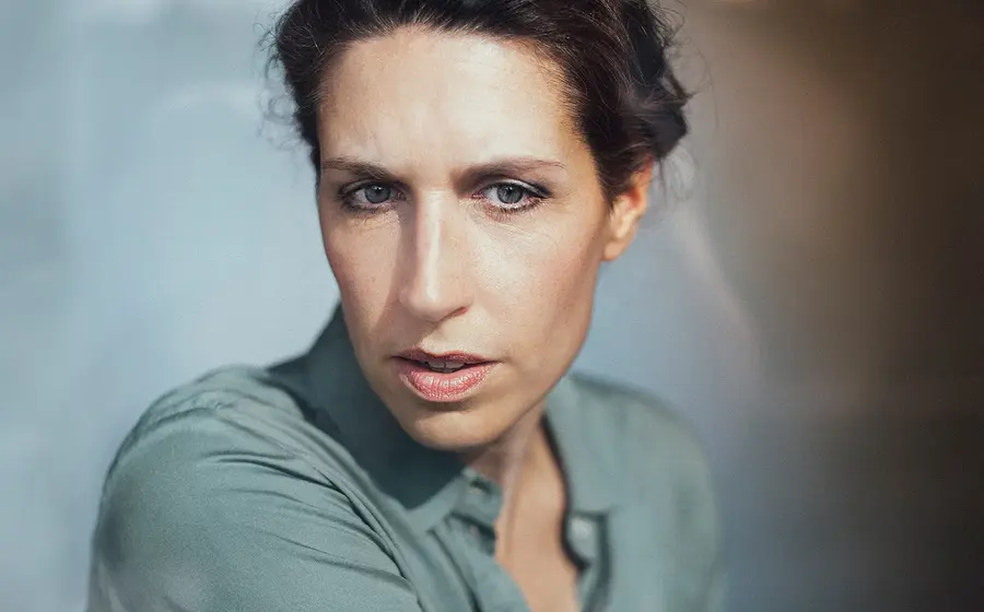 Portrait von Christine Prayon in türkiser Bluse vor hellblau-hellbraunem Hintergrund mit Farbverlauf