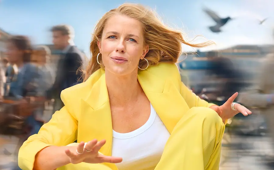Christine Eixenberger in leuchtend gelbem Hosenanzug sitzt in dynamischer Pose im Freien. Im Hintergrund ziehen verschwommene Passantinnen und Passanten vorbei, während Wind durch die Haare weht und Vögel am Himmel zu sehen sind.