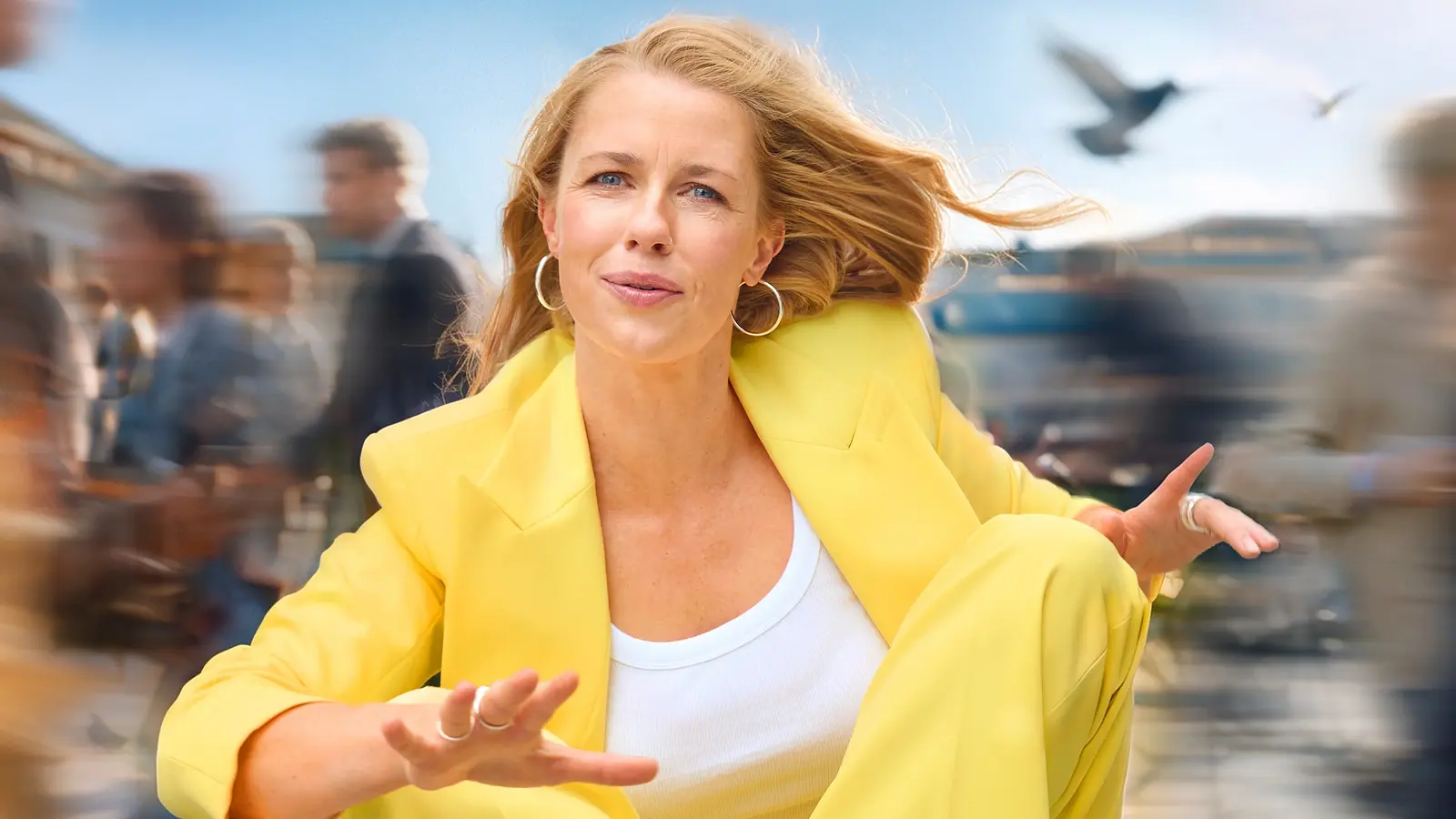 Christine Eixenberger in leuchtend gelbem Hosenanzug sitzt in dynamischer Pose im Freien. Im Hintergrund ziehen verschwommene Passantinnen und Passanten vorbei, während Wind durch die Haare weht und Vögel am Himmel zu sehen sind.