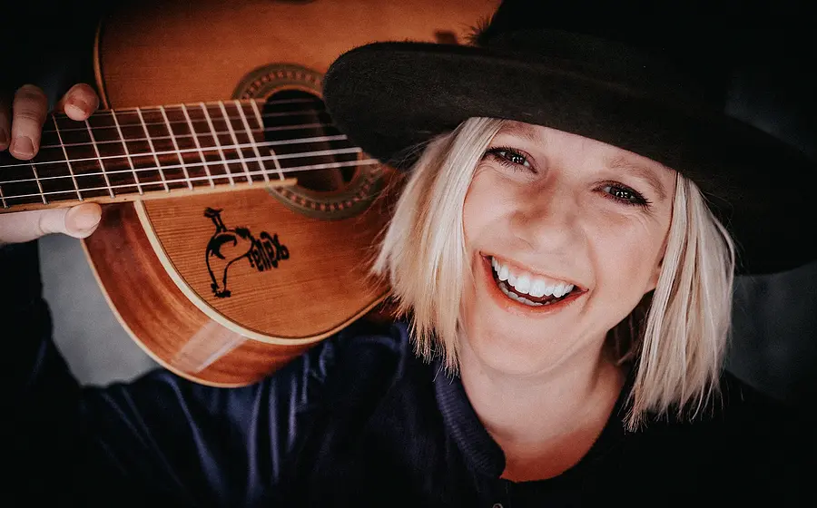 Claudia Koreck mit hellem, kurzem Haar und schwarzem Hut hält eine Gitarre auf der Schulter. Nahaufnahme vor dunklem Hintergrund.