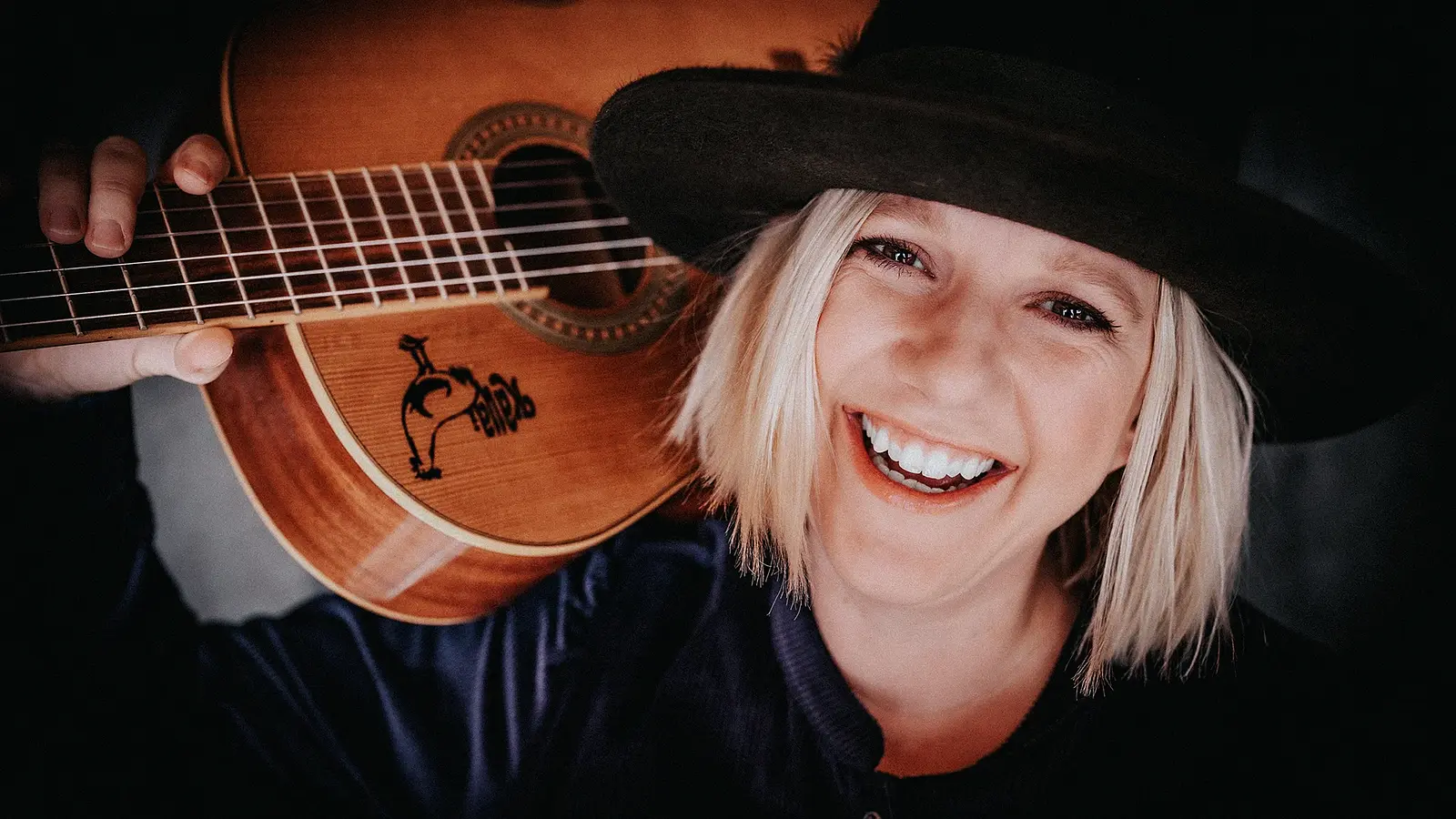 Claudia Koreck mit hellem, kurzem Haar und schwarzem Hut hält eine Gitarre auf der Schulter. Nahaufnahme vor dunklem Hintergrund.