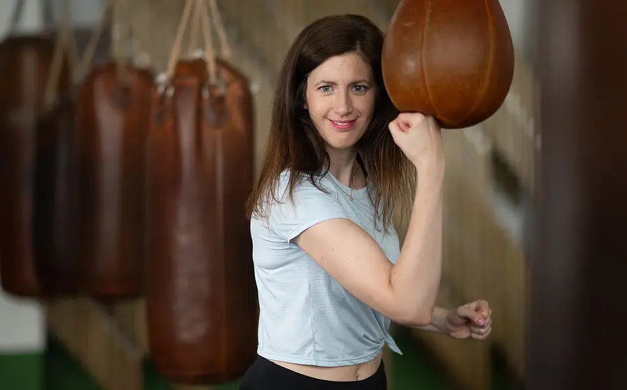 Frau trainiert mit einem Boxsack in einem Fitnessstudio, lächelnd in sportlicher Kleidung, mit mehreren Sandsäcken im Hintergrund.