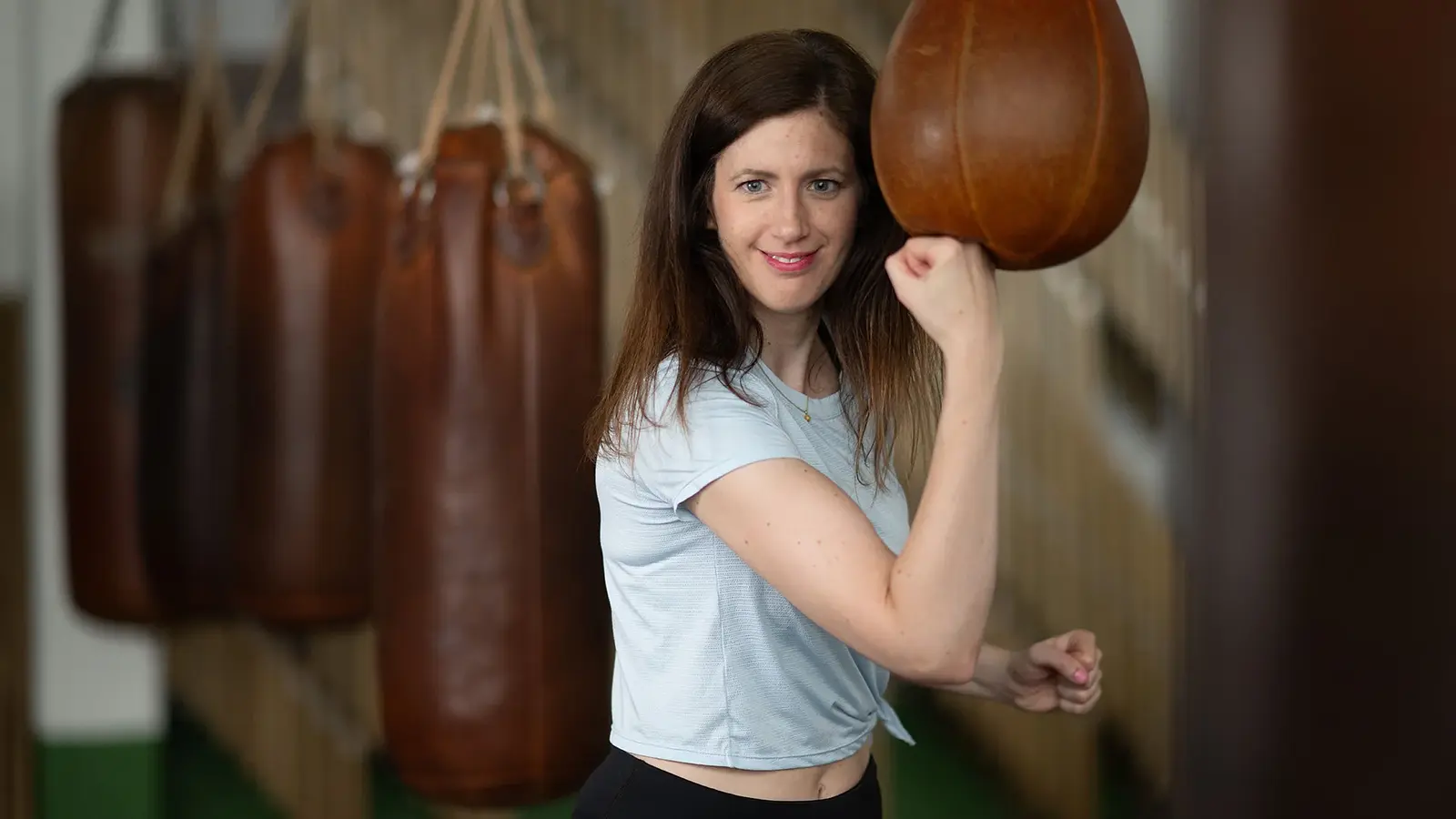 Frau trainiert mit einem Boxsack in einem Fitnessstudio, lächelnd in sportlicher Kleidung, mit mehreren Sandsäcken im Hintergrund.