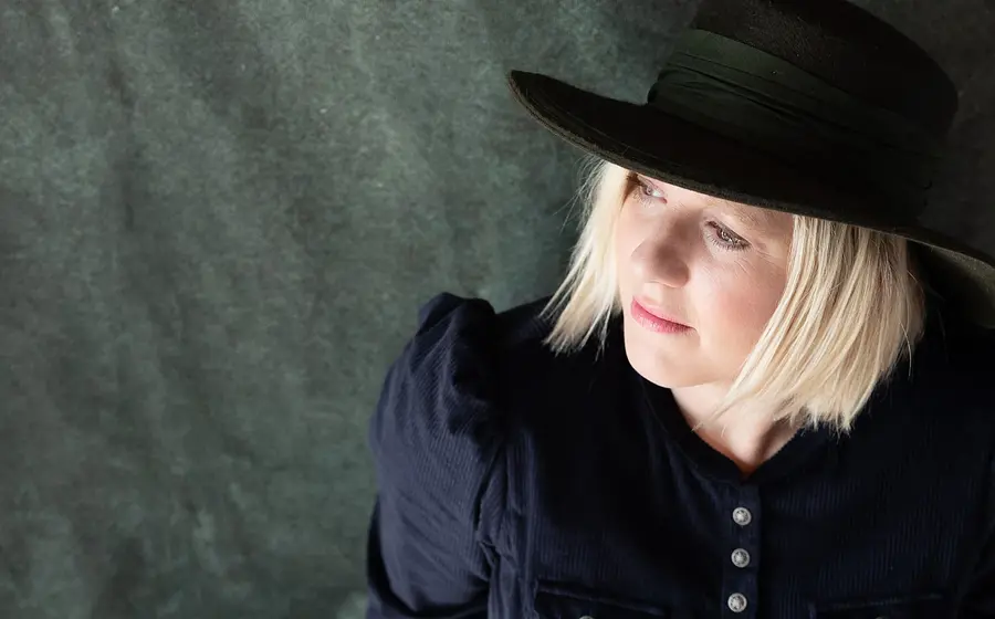 Claudia Koreck mit schwarzem Hut und dunkelblauem Oberteil blickt zur Seite, vor grau marmoriertem Hintergrund