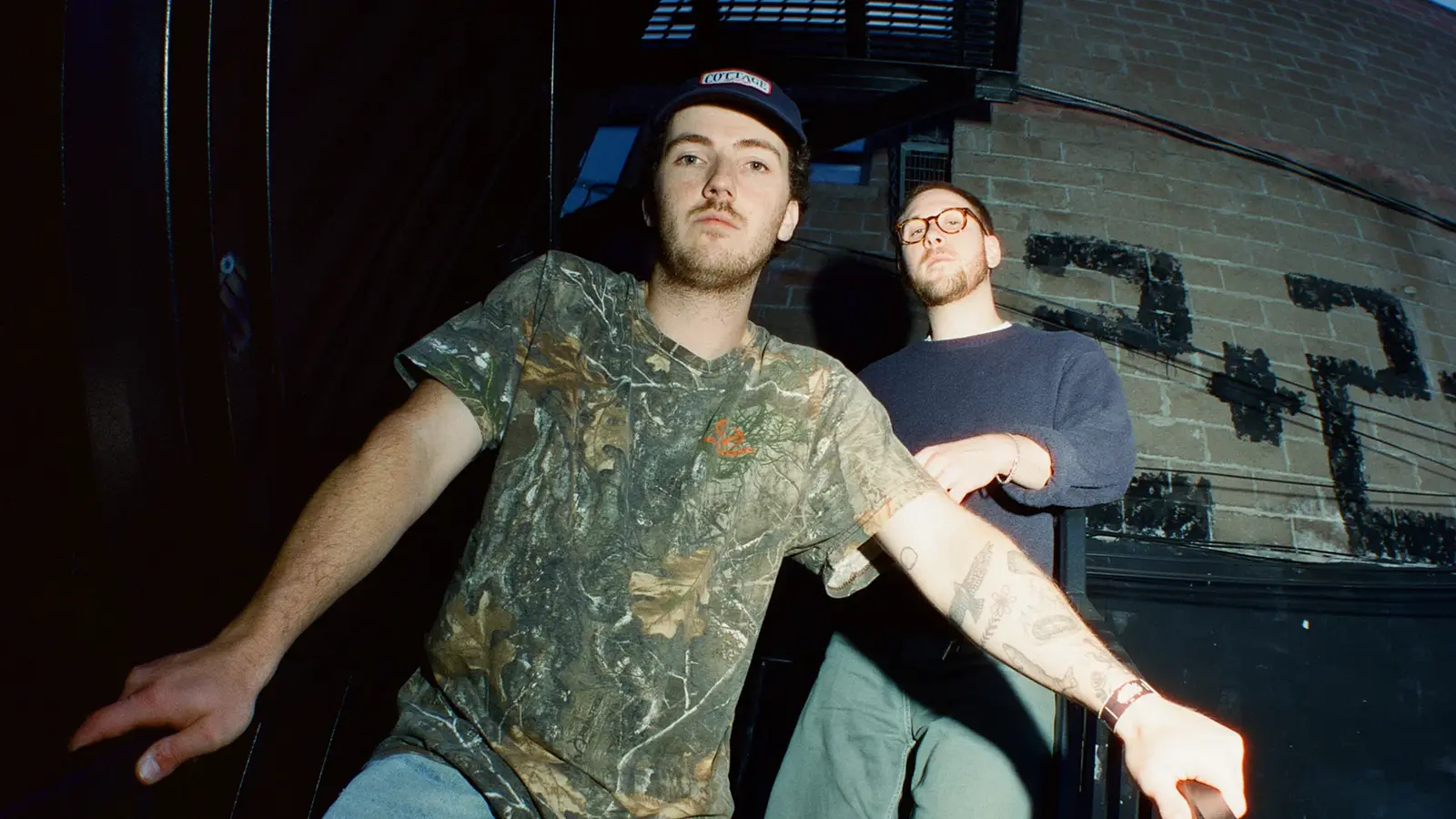 Bandfoto von Cleopatrick mit zwei Bandmitgliedern vor einer Backsteinwand im Außenbereich, eine Person in Tarnmuster-T-Shirt und Kappe im Vordergrund, die andere mit Brille und dunklem Pullover dahinter, aufgenommen aus tiefer Perspektive bei direktem Blitzlicht.