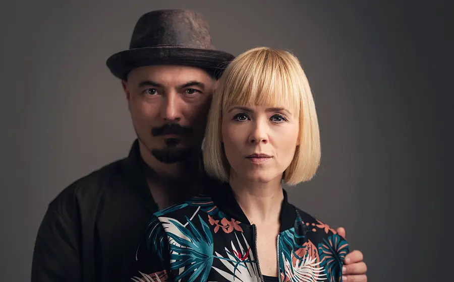 Ein Portrait vom Duo Carolin No vor einem grauen Hintergrund eine Person mit Hut steht hinter einer Person mit blondem Bob und gemusterter Jacke.