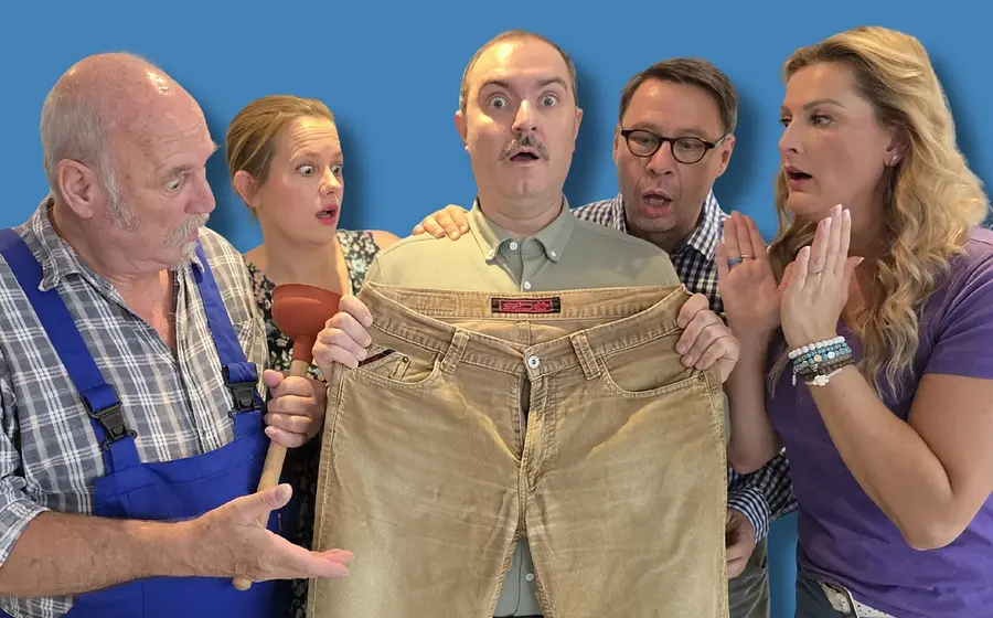 Fünf Darsteller stehen eng zusammen, eine Person hält eine beige Hose hoch, während die anderen darauf schauen, vor blauem Hintergrund