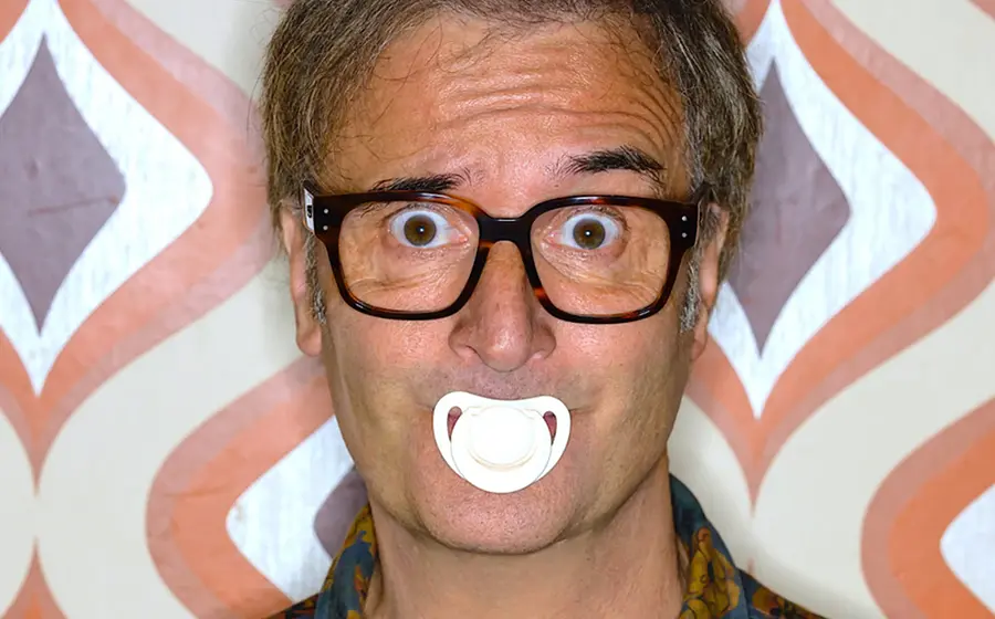 Ein Portrait von Chris Boettcher mit enem weißen Schnuller im Mund vor einer Tapete mit Muster in braun, beige und orange