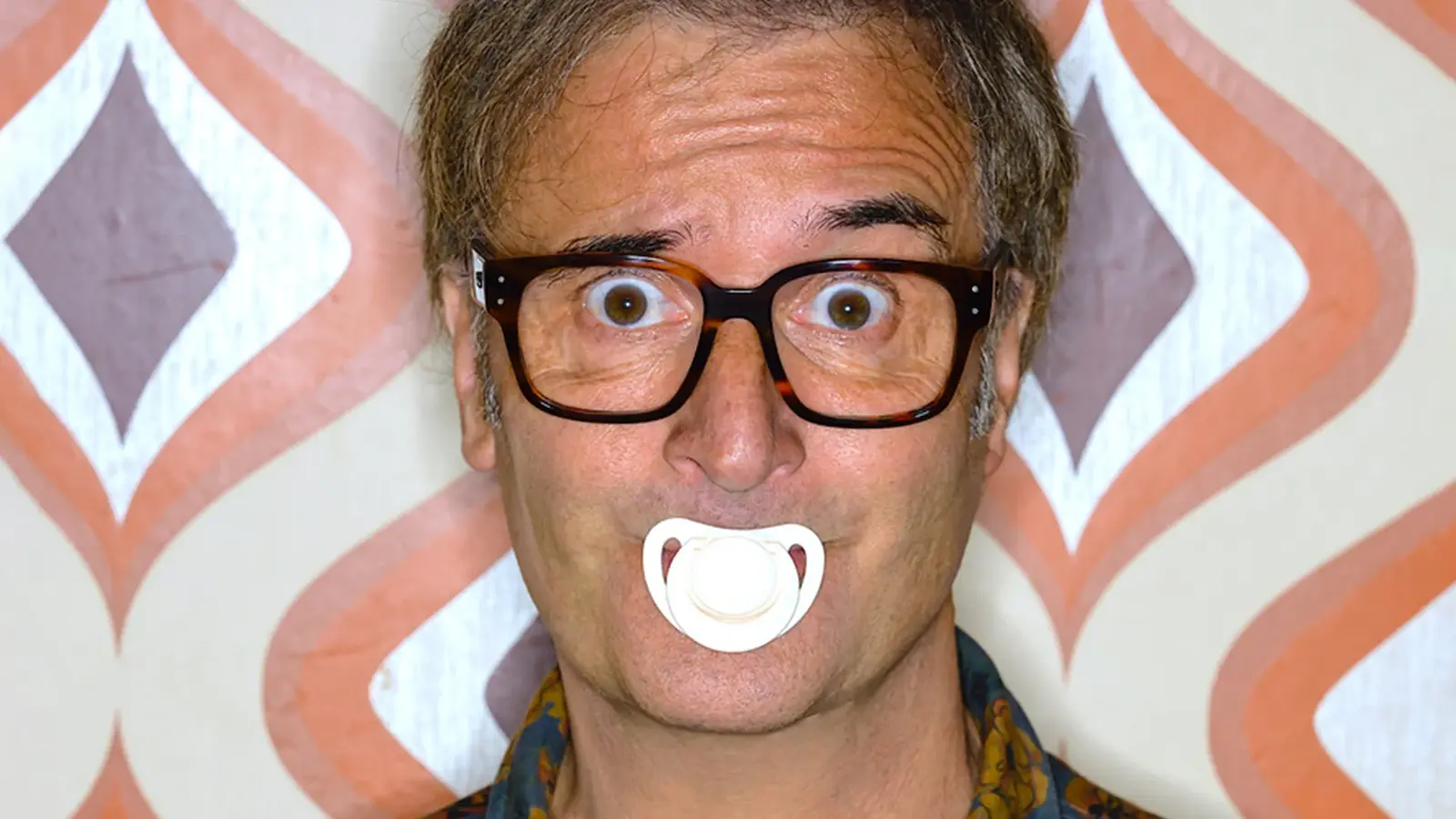 Ein Portrait von Chris Boettcher mit enem weißen Schnuller im Mund vor einer Tapete mit Muster in braun, beige und orange