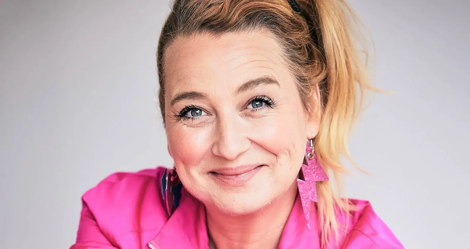 Ein Portrait von Constanze Lindner mit pinkem Oberteil und pinken Blitzförmigen Ohrringen vor weißem Hintergrund