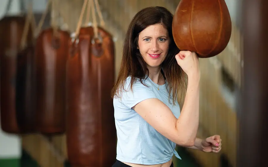 Claudia Pichler im hellblauen T Shirt, die mit erhobenem Arm einen Boxsack schlägt, in Trainingsraum mit mehreren braunen Boxsäcken im Hintergrund