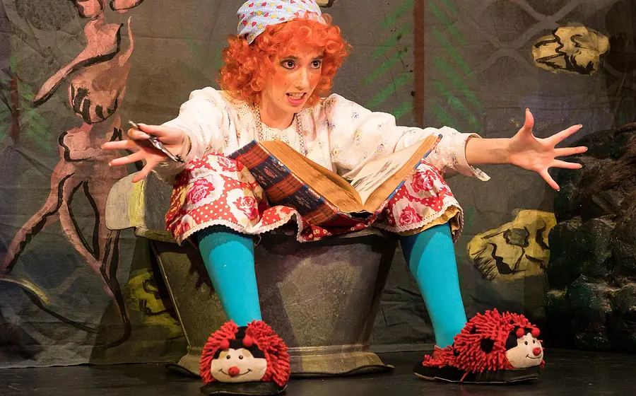 Foto für das Event Die kleine Hexe, mit einer Frau, die spielt und verkleidet ist, in einem Würfel sitzt und ein Buch sowie Marienkäfer‑Hausschuhe hat.