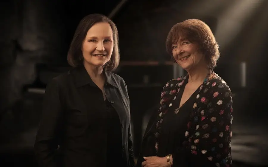 Eva Barbarion und Ulrike Zöller in dunkler Kleidung stehen beisammen in dunklem gedämpften Licht und lächeln in die Kamera