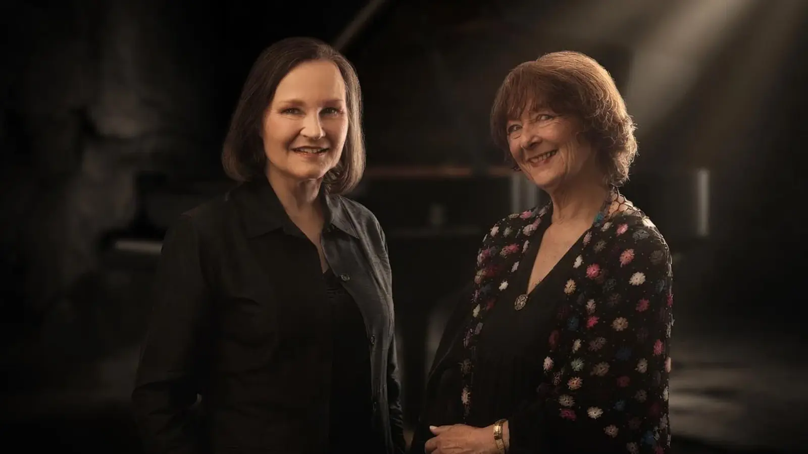 Eva Barbarion und Ulrike Zöller in dunkler Kleidung stehen beisammen in dunklem gedämpften Licht und lächeln in die Kamera
