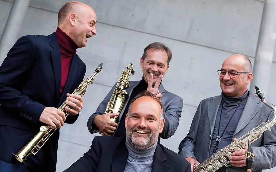 Vier Musike in dunklen Anzügen, drei davon mit Blsinstrumenten , blicken lachend in die Kamera, der Hintergrund eine graue Betonwand
