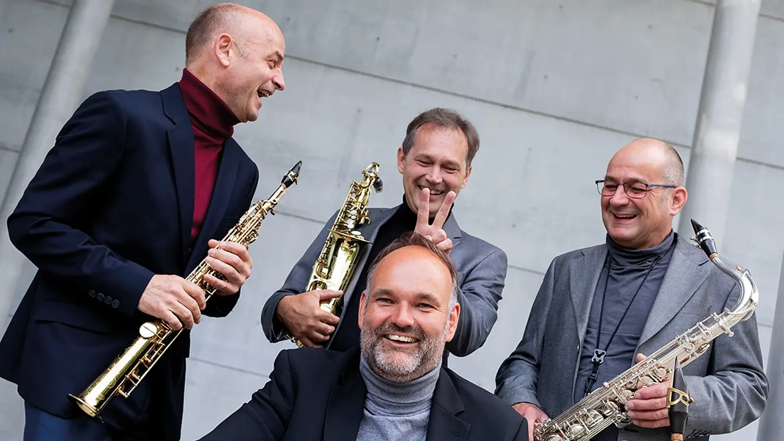 Vier Musike in dunklen Anzügen, drei davon mit Blsinstrumenten , blicken lachend in die Kamera, der Hintergrund eine graue Betonwand
