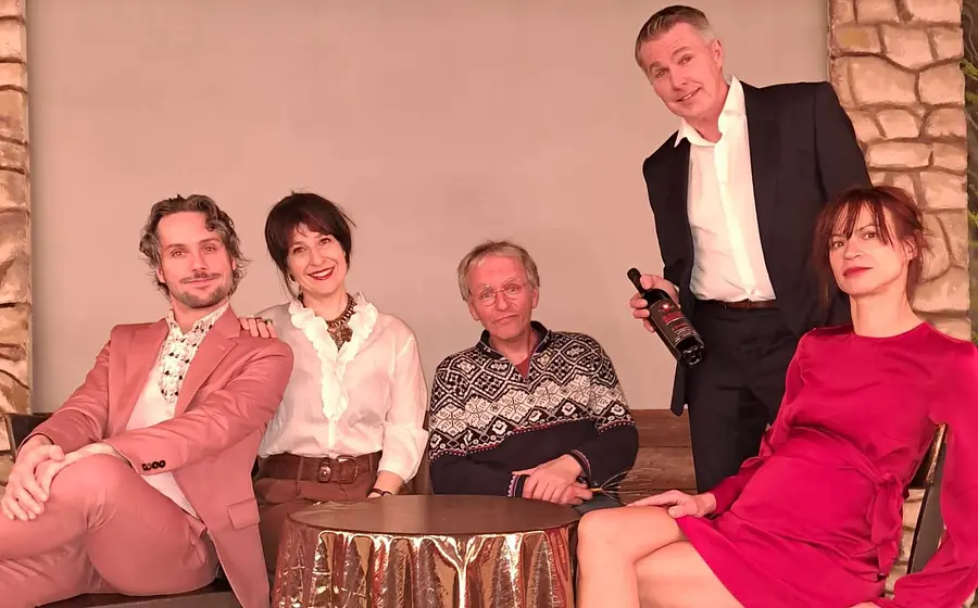 Die Schauspieler des Stückes der Vorname um einen Tisch herum und sie lächeln in die Kamera