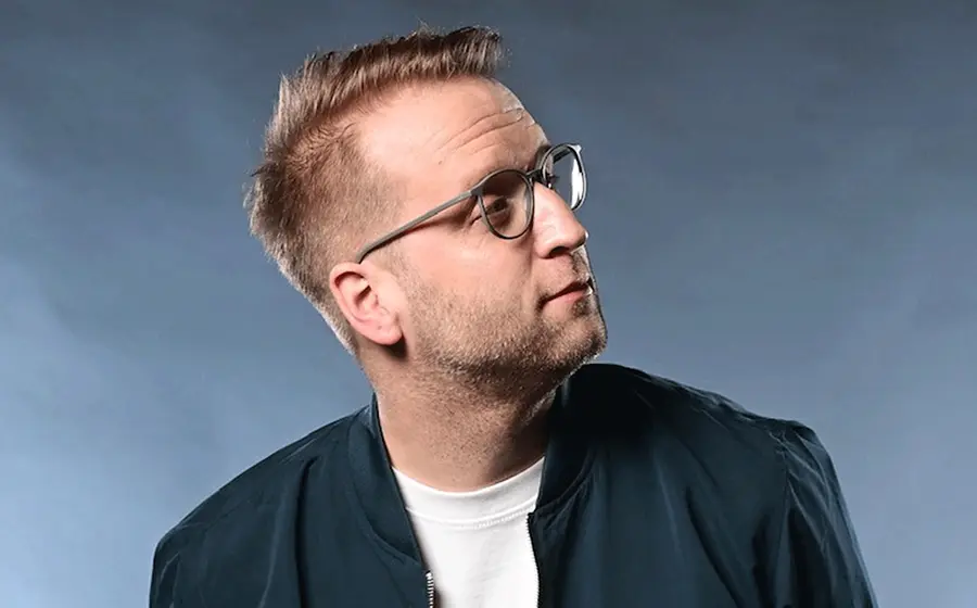 Benni Stark im Porträt mit Brille und dunkler Jacke vor blauem Hintergrund, den Blick zur Seite gerichtet.