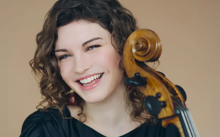 Portrait Julia Hagen mit dem Hals eines Violoncello in schwarzer Kleidung lachend vor beigem Hintergrund