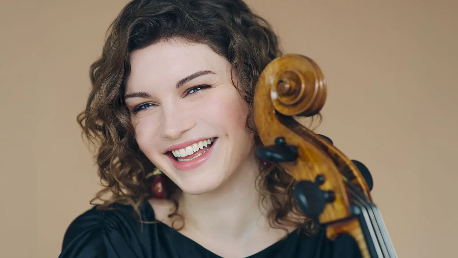 Portrait Julia Hagen mit dem Hals eines Violoncello in schwarzer Kleidung lachend vor beigem Hintergrund