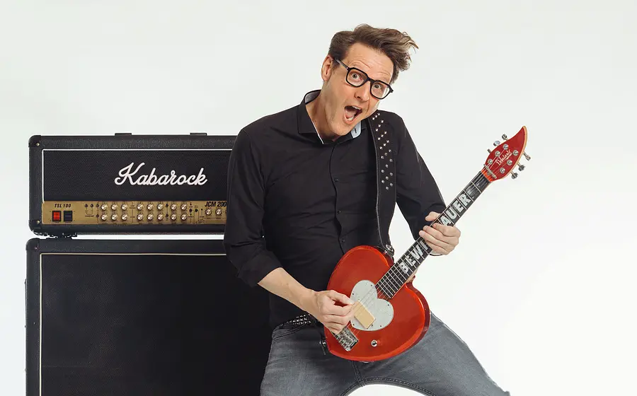 Bewie Bauer mit Brille auf spielt eine rote E Gitarre vor einem Verstärker mit der Aufschrift Kabarock und posiert in dynamischer Haltung vor weißem Hintergrund