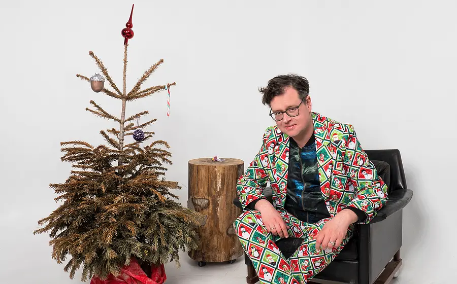 Der Comedian sitzt mit auffälligem Weihnachtsanzug in einem schwarzen Sessel neben einem vertrockneten Weihnachtsbaum mit spärlichem Baumschmuck vor weißem Hintergrund.