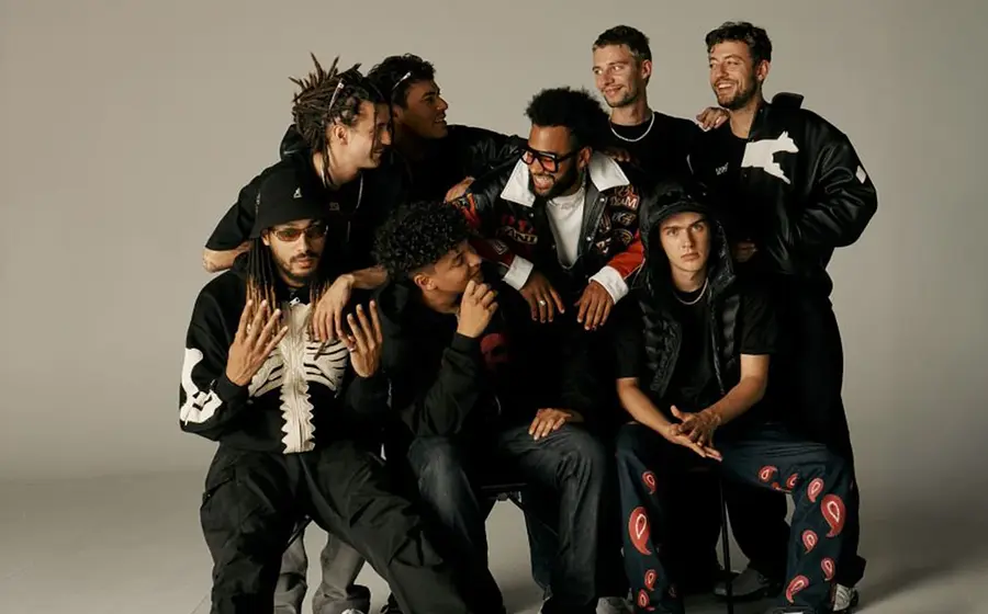 Bandfoto von BHZ mit acht Bandmitgliedern in dunkler Freizeitkleidung vor hellem Studiohintergrund, einige sitzend, andere stehend, mit Kappen, Brillen und auffälligen Jacken in lockerer Gruppenanordnung