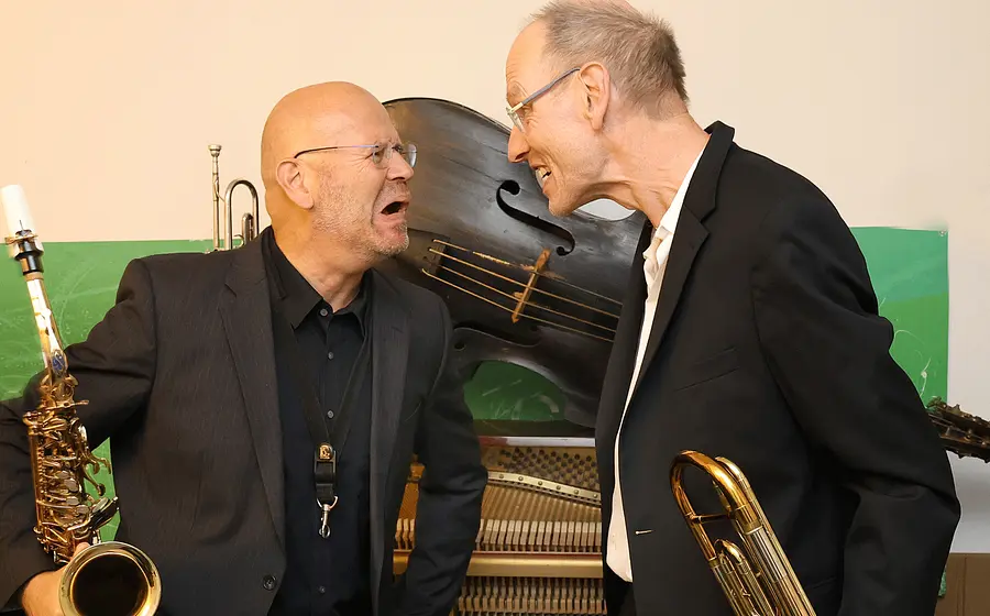 Zwei Musiker stehen sich mit Blasinstrumenten gegenüber und posieren vor einem Klavier sowie einem großen Kontrabass. Eine Person hält ein Saxofon, die andere ein tiefes Blechblasinstrument, während beide einander zugewandt sind.