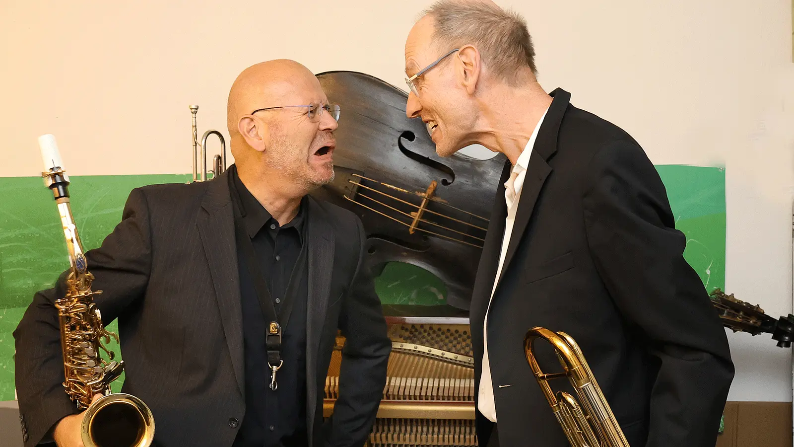 Zwei Musiker stehen sich mit Blasinstrumenten gegenüber und posieren vor einem Klavier sowie einem großen Kontrabass. Eine Person hält ein Saxofon, die andere ein tiefes Blechblasinstrument, während beide einander zugewandt sind.