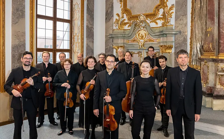 Kammerorchester mit mehreren Musikerinnen und Musikern in schwarzer Konzertkleidung, die Streichinstrumente halten, posiert in einem historischen, reich verzierten Saal mit großen Fenstern und goldenen Ornamenten.